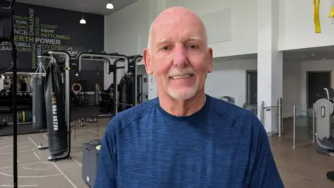 John is wearing a blue tshirt and is stood in a leisure centre with exercise equipment behind him.