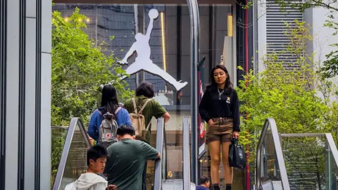 Getty Images Shoppers ride escalators near a Jordan Brand retail store, with the iconic Jumpman logo visible on the storefront, surrounded by urban greenery and modern commercial architecture
